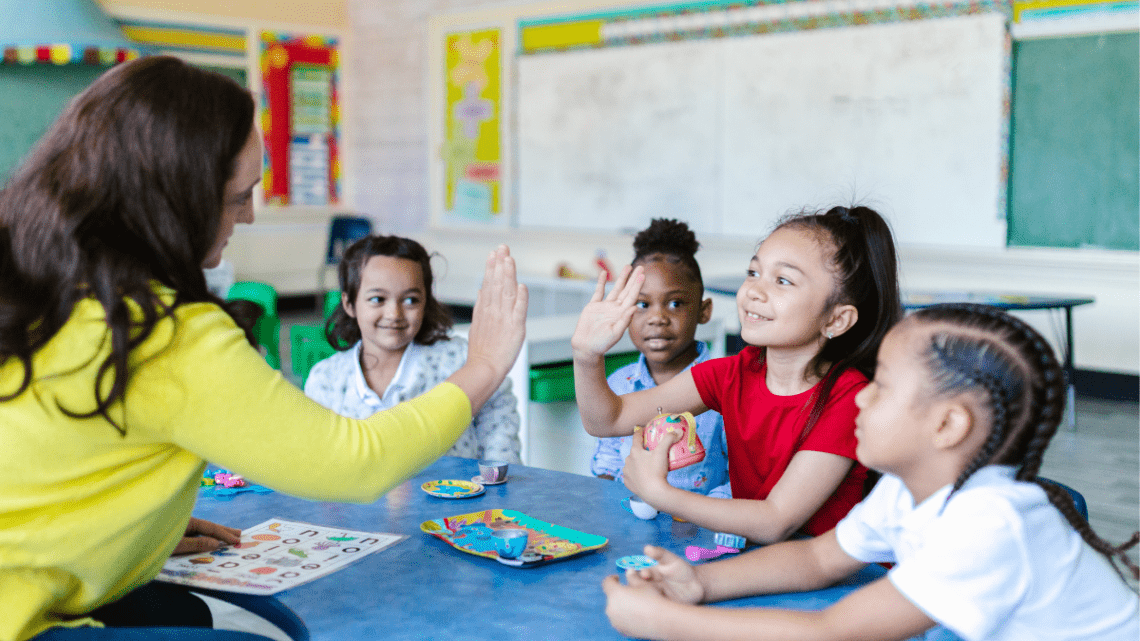 inclusive classroom of kids roughly ages 6 or 7 and one is high fiving her teacher. The teacher wears a yellow short and has long black hair.