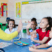 inclusive classroom of kids roughly ages 6 or 7 and one is high fiving her teacher. The teacher wears a yellow short and has long black hair.