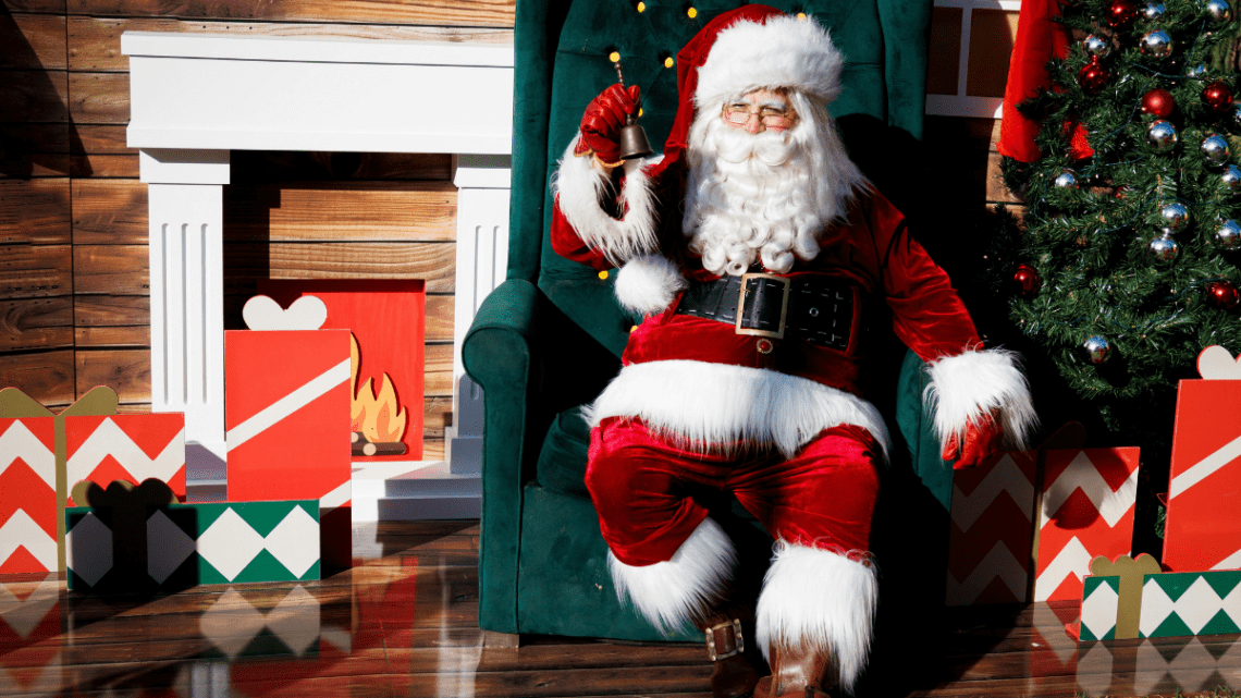 santa in full costume sitting on a chair in front of a fireplace with gifts all over the floor wrapped prettily in bright coloured paper