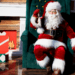 santa in full costume sitting on a chair in front of a fireplace with gifts all over the floor wrapped prettily in bright coloured paper