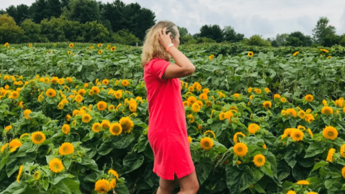 woman standing in a field of sunflowers on a sunny day in august. She is wearing a coral coloured dress and looking out to the horizon while holding back her blonde-ish hair in the wind
