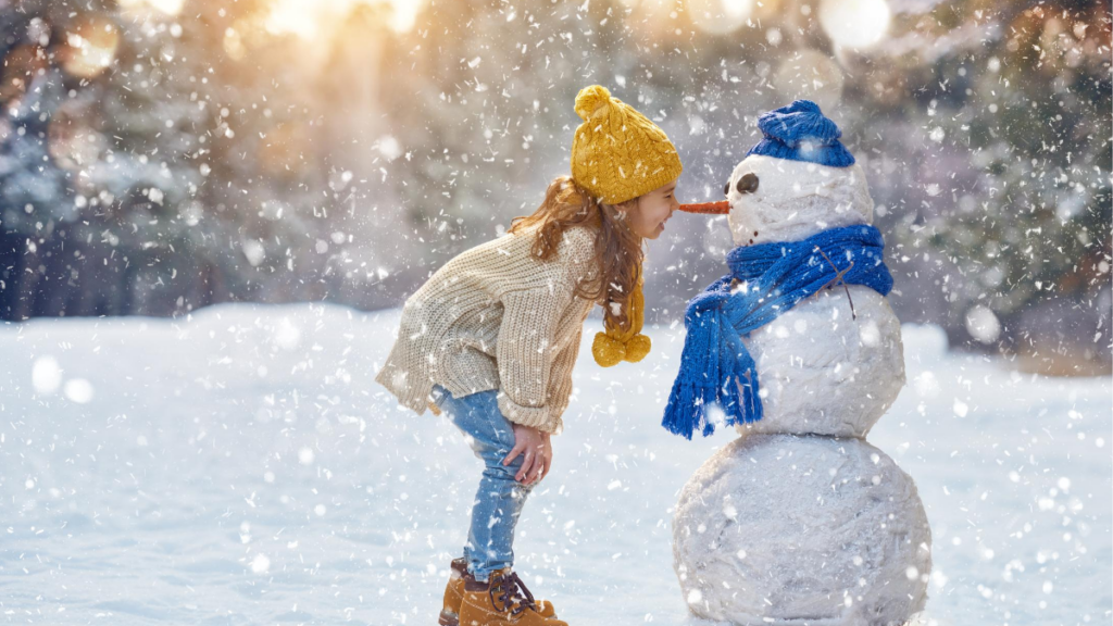 young girl standing in falling snow looking at her snowman with carrot nose and blue hat and scarf