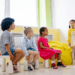 several kids sitting in a circle watching a young kid acting like a teacher and reading to them