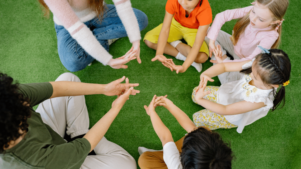 preschool kids in a circle sitting on a green carpet that looks like lawn stretching their hands