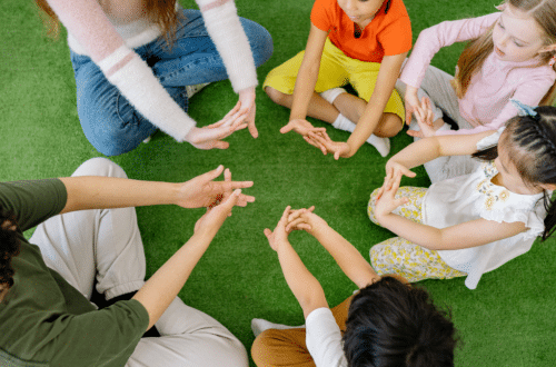 preschool kids in a circle sitting on a green carpet that looks like lawn stretching their hands