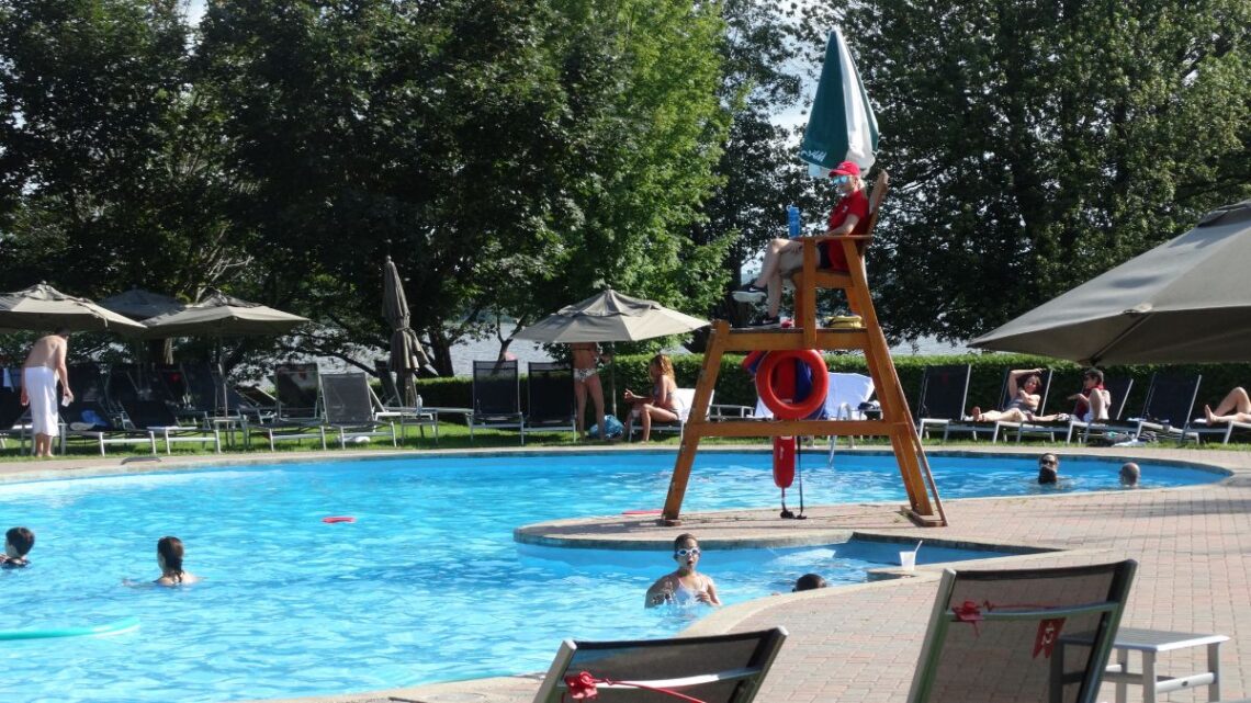 big public pool deck at a resort with colourful umbrellas and a life guard chair. several people of all ages are in the pool and it is a sunny day