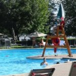 big public pool deck at a resort with colourful umbrellas and a life guard chair. several people of all ages are in the pool and it is a sunny day