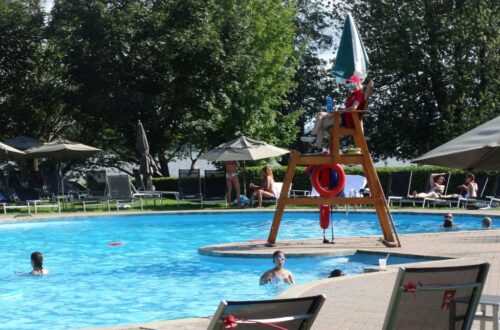 big public pool deck at a resort with colourful umbrellas and a life guard chair. several people of all ages are in the pool and it is a sunny day