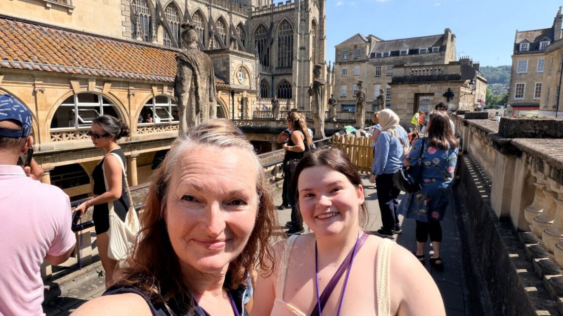 roman baths and two women on the top level of a Georgian building that appears open below with ancient city in background and green water in the baths below