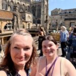 roman baths and two women on the top level of a Georgian building that appears open below with ancient city in background and green water in the baths below