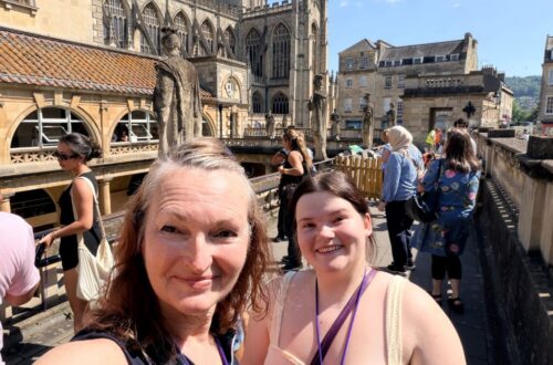 roman baths and two women on the top level of a Georgian building that appears open below with ancient city in background and green water in the baths below
