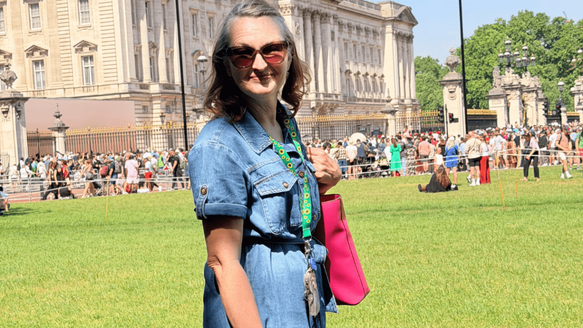 woman in a denim dress standing in front of Buckingham palace wearing the sunflower lanyard and smiling with bright pink tote bag purse