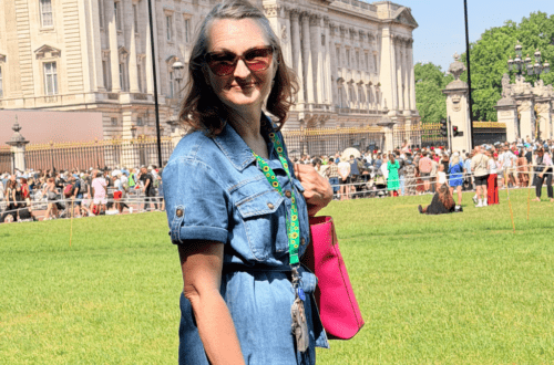 woman in a denim dress standing in front of Buckingham palace wearing the sunflower lanyard and smiling with bright pink tote bag purse