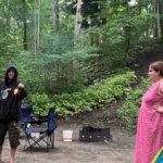 sable beach camping at provincial park trees in the background and two young adults in front of a tent. One is in black and covered and getting ready to start a fire