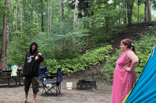 sable beach camping at provincial park trees in the background and two young adults in front of a tent. One is in black and covered and getting ready to start a fire
