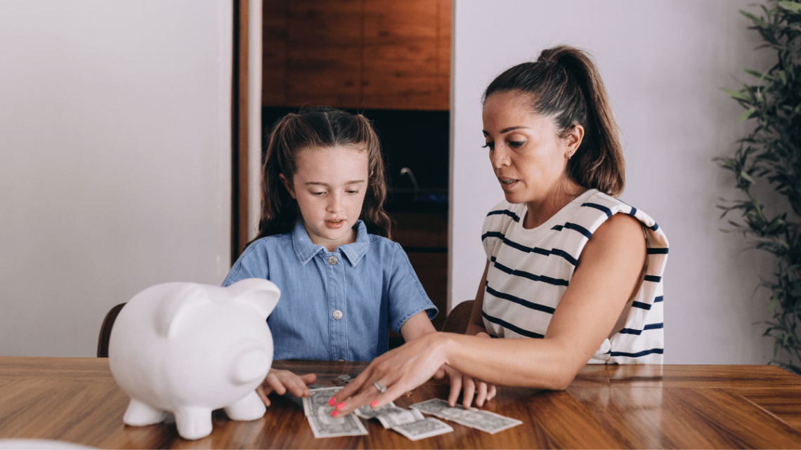 little girl counting dollar bills beside a white piggy bank and her mom is helping her looking intensely at the dollar bills. Closet behind them and they are dressed in light clothing. Girl has denim top on and her mom has a short sleeved white and navy striped top. Navy stripes are thin
