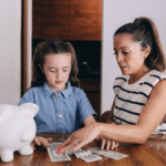 little girl counting dollar bills beside a white piggy bank and her mom is helping her looking intensely at the dollar bills. Closet behind them and they are dressed in light clothing. Girl has denim top on and her mom has a short sleeved white and navy striped top. Navy stripes are thin