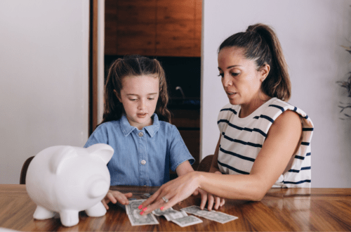 little girl counting dollar bills beside a white piggy bank and her mom is helping her looking intensely at the dollar bills. Closet behind them and they are dressed in light clothing. Girl has denim top on and her mom has a short sleeved white and navy striped top. Navy stripes are thin