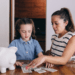 little girl counting dollar bills beside a white piggy bank and her mom is helping her looking intensely at the dollar bills. Closet behind them and they are dressed in light clothing. Girl has denim top on and her mom has a short sleeved white and navy striped top. Navy stripes are thin