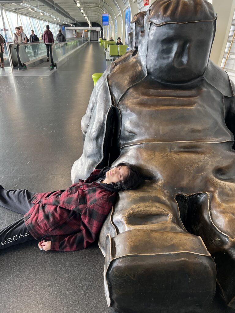 19-year-old young person with red plaid jacket lying on the floor of the airport in Amsterdam. Head propped up on a bronze statue. The youth is wearing black Spyder track pants and looks pale. Walking track through airport is behind them. It is fall based on clothing and the tunnel like room is well lit.