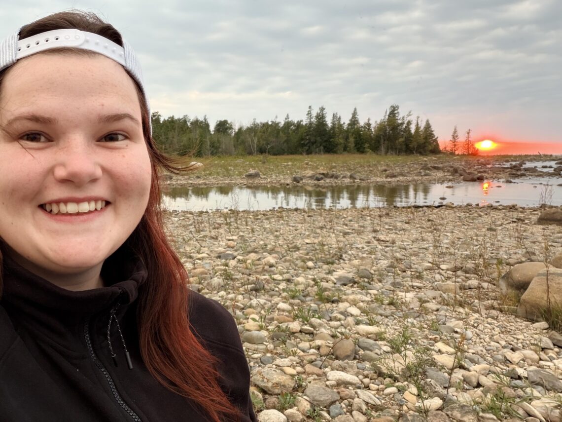young girl with red hair looking at the camera and smiling with black sweater on and rocks across the horizon as the sunsets behind her on a Rocky point and green evergreens across the horizon