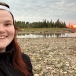 young girl with red hair looking at the camera and smiling with black sweater on and rocks across the horizon as the sunsets behind her on a Rocky point and green evergreens across the horizon