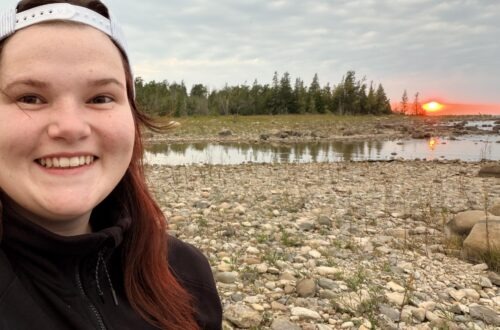 young girl with red hair looking at the camera and smiling with black sweater on and rocks across the horizon as the sunsets behind her on a Rocky point and green evergreens across the horizon