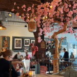 Inside a colourful restaurant in Alkmaar Netherlands. A woman is seated at a table in the foreground. There are cherry blossoms that are pink hanging from the ceiling off of a fake tree with twinkling lights and brown brick walls that make it look cozy. In behind a server is waiting on a table and the restaurant is about half full but not overly crowded. The server appears to have Down syndrome