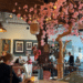 Inside a colourful restaurant in Alkmaar Netherlands. A woman is seated at a table in the foreground. There are cherry blossoms that are pink hanging from the ceiling off of a fake tree with twinkling lights and brown brick walls that make it look cozy. In behind a server is waiting on a table and the restaurant is about half full but not overly crowded. The server appears to have Down syndrome