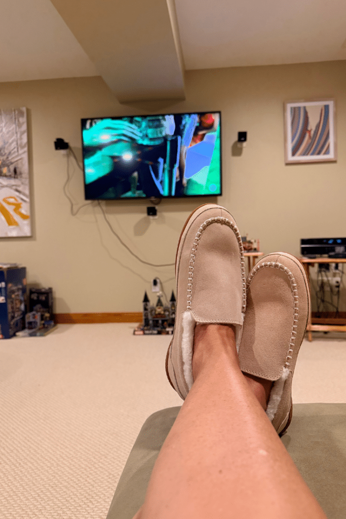 bare legs up on a lounge chair in the basement. light beige berber carpet on the ground and moccasin style sand coloured slippers on feet. Tv is on and streaming in the background. It is a big screen tv hanging on the wall.